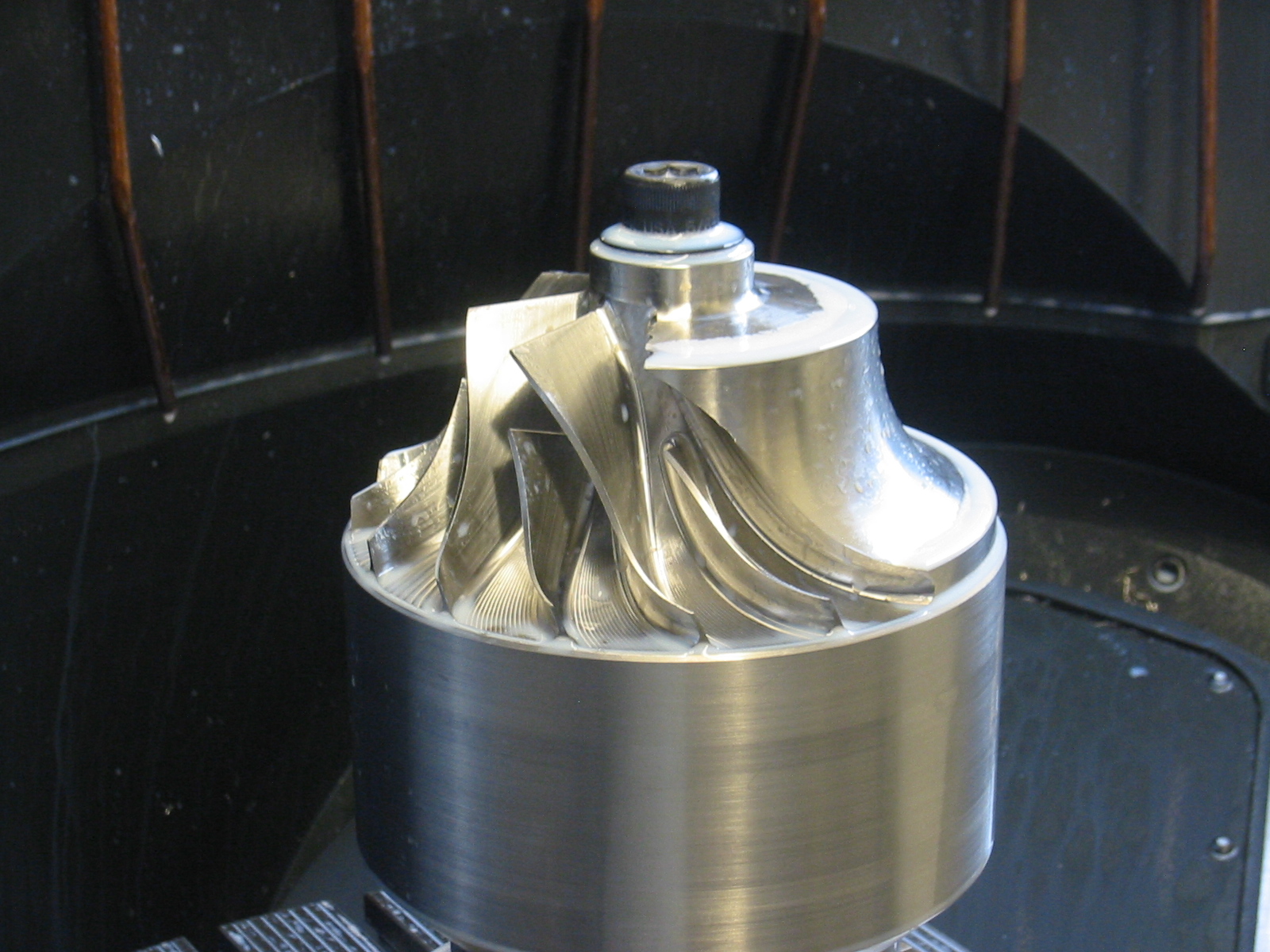 A titanium impeller test part with curved machined blades mounted inside a machine enclosure, with no people visible.